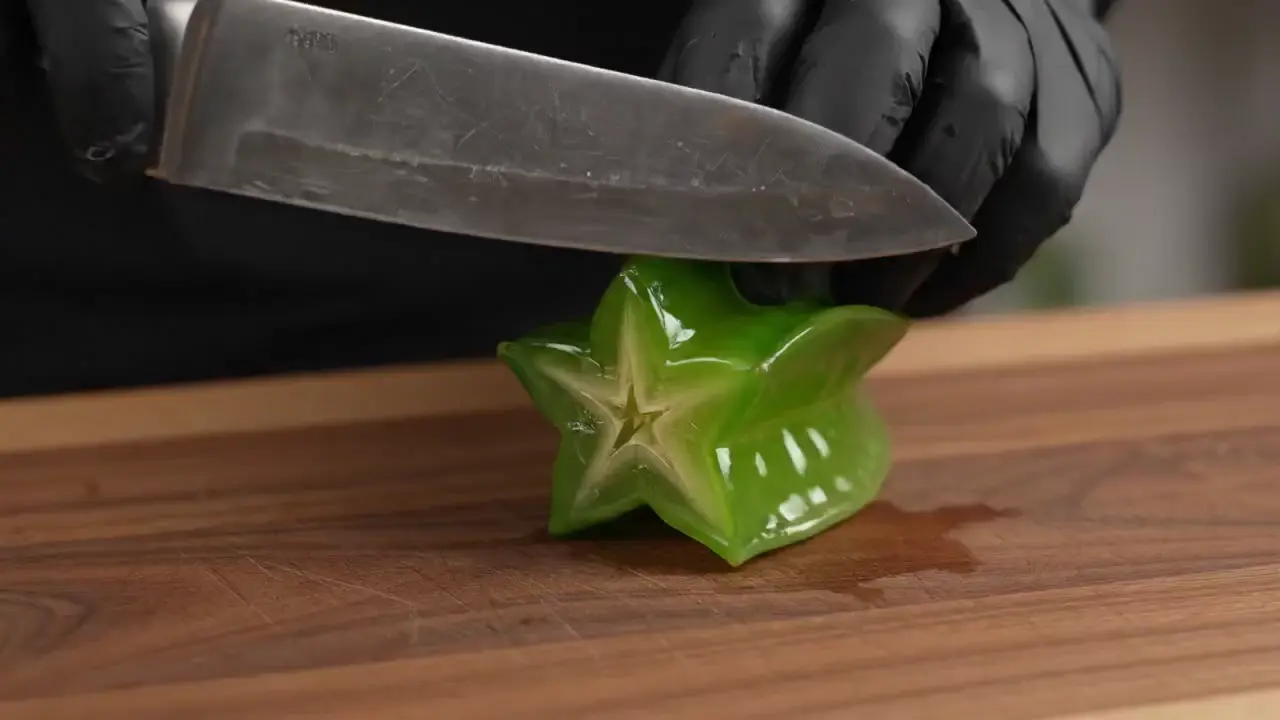 Gloved hand slicing a green glass starfruit with a chef's knife on a wooden cutting board